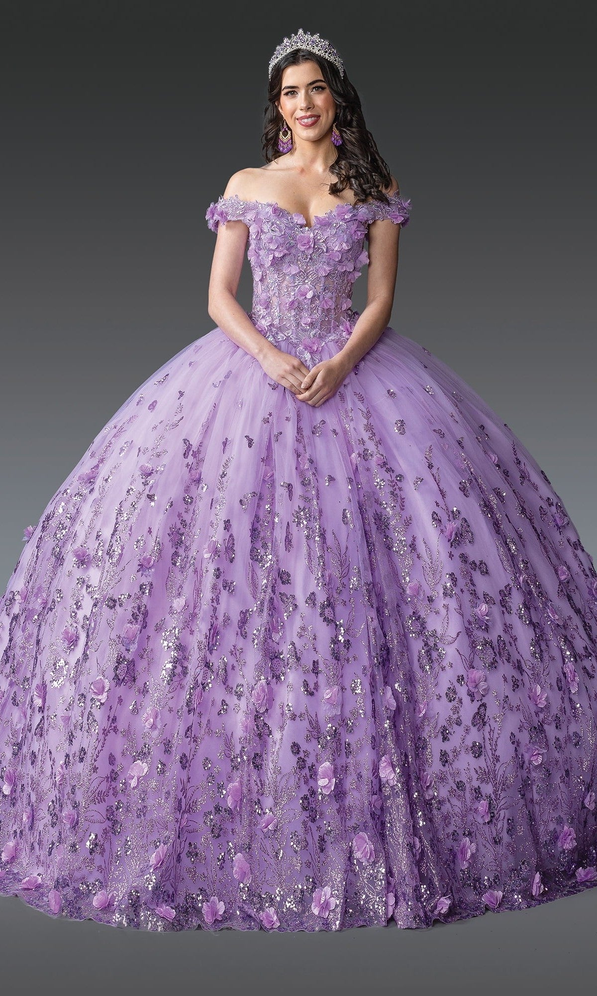 A woman wearing the Quinceanera Dress 1908 by Dancing Queen in lilac, featuring delicate 3D floral appliques and off-the-shoulder sleeves, accessorized with a jeweled tiara and earrings, stands against a plain gray background.